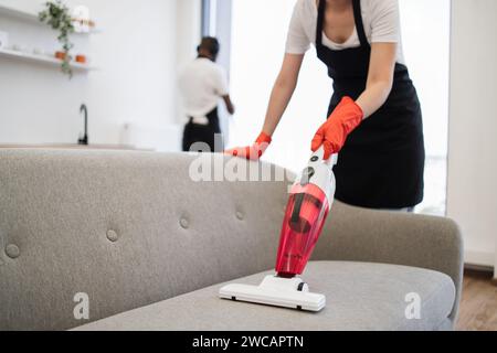 Vue recadrée des mains de jeune nettoyeur féminin multiethnique aspirant canapé avec aspirateur sans fil portable. Heureux souriant employés de nettoyage co Banque D'Images