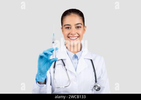 Une femme médecin joyeuse dans un manteau blanc et des gants bleus affiche en toute confiance une seringue Banque D'Images