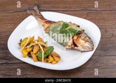 Poisson dorado cuit au four dans des feuilles de bambou, pommes de terre cuites avec des pousses de bambou sur une assiette blanche. Cuisine maison. Banque D'Images