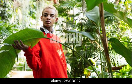 Un jeune homme aux cheveux blonds dans un pull rouge et une chemise blanche pose dans un jardin botanique verdoyant. Banque D'Images