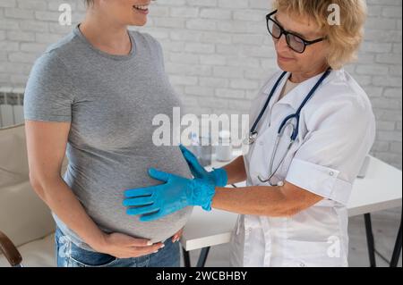 Femme enceinte consultant un médecin. Gynécologue caucasienne âgée tient les mains sur le ventre d'une patiente enceinte. Banque D'Images