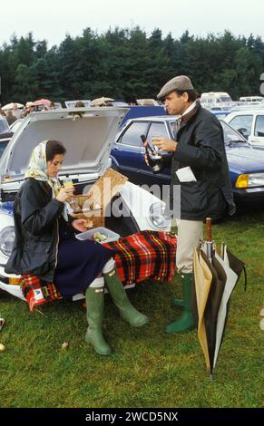 Couple Sloane Ranger profitant d'un pique-nique en plein air depuis leur cabriolet Porsche 911 SC Cabriolet. Elle est sur un Bucks Fizz, mais fait avec Henkell Trocken, un vin blanc mousseux allemand, pendant qu'il le prend soigné. Ils sont tous les deux habillés comme le dicte la mode pour l'occasion dans de nouvelles bottes Hunter Wellington vertes non boueuses et ses vestes bleues Burberry cirées, absolument en vogue à l'époque. Elle a enfilé un foulard Hermès, alors qu'il est dans une casquette plate en tweed. Windsor, Berkshire, Angleterre juin 1985 1980s HOMER SYKES Banque D'Images