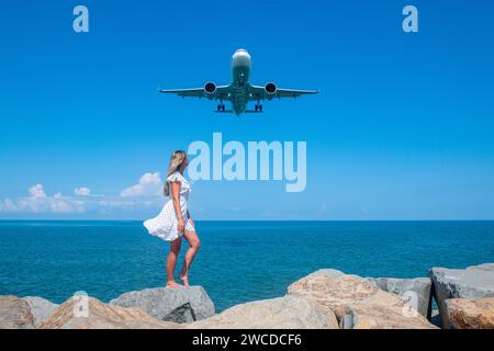 Sérénité aérienne : fille en robe blanche sur pierres, avion s'élevant au-dessus de la mer Bleue Banque D'Images