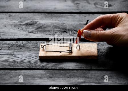 Un piège à souris avec un morceau de saucisse se tient sur un plancher noir en bois dans la maison, et la main d'une femme met la saucisse dans le piège à souris, attrapant des souris Banque D'Images