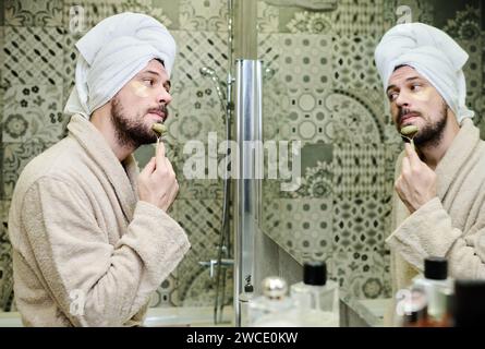 Jeune homme en peignoir ayant procédé de massage facial avec rouleau de pierre de jade après la douche du matin tout en se tenant devant le miroir Banque D'Images