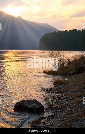 Paysage de coucher de soleil de l'Altaï du soir avec la rivière Katun et des rochers au premier plan et des montagnes avec forêt sur la rive lointaine. Le Soleil se couche derrière la montagne Banque D'Images