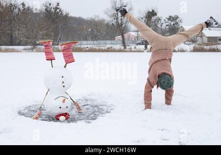 bonhomme de neige drôle se tient à l'envers sur un lac enneigé gelé. une jeune femme méconnaissable à proximité fait un transfert. loisirs actifs saisonniers. Vacances en Banque D'Images