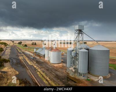 Vue aérienne de silos à grains le long d'une ligne de chemin de fer rurale avec des nuages sombres et de la pluie au loin à Moolort dans le Victoria central, Australie. Banque D'Images