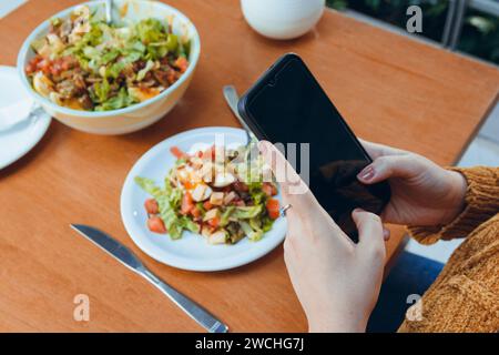 Gros plan des mains féminines caucasiennes d'une femme méconnaissable assise tenant le téléphone envoyant un message pendant le déjeuner, avec une salade sur la table en arrière-plan. copier Banque D'Images