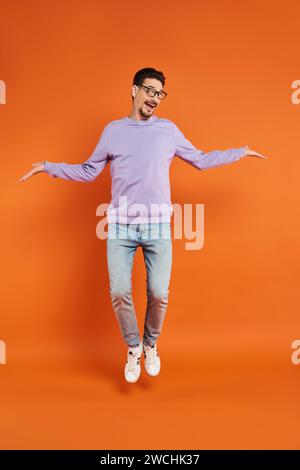 homme joyeux dans les lunettes et pull violet lévitant sur fond orange, volant dans les airs Banque D'Images
