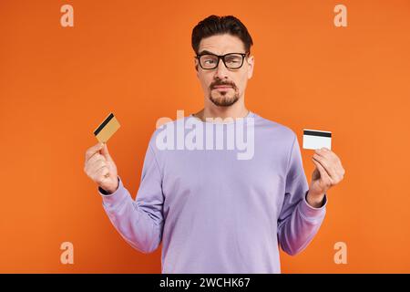 homme sceptique en lunettes et pull violet comparant deux cartes de crédit sur fond orange Banque D'Images