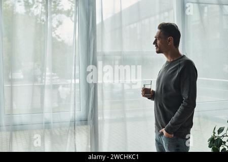 homme barbu réfléchi debout avec la main dans la poche, regardant la fenêtre et tenant un verre d'eau Banque D'Images