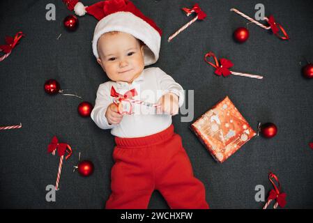 Joyeux bébé garçon heureux dans le chapeau du Père Noël souriant parmi les cannes de bonbons de Noël, boules rouges et boîte-cadeau Banque D'Images