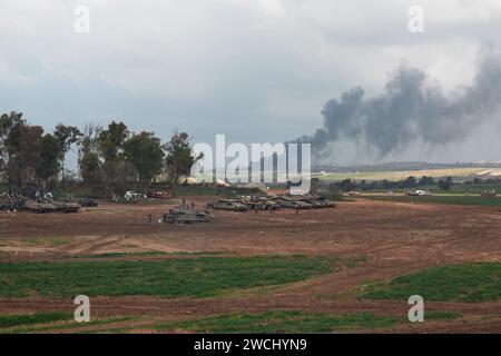 Un char militaire israélien roule à côté de chars à une position près de la frontière avec la bande de Gaza alors que de la fumée massive gonfle après le bombardement israélien dans le centre de Gaza au milieu des combats continus entre Israël et le groupe militant Hamas le 14 janvier 2024 à la frontière de Gaza, Israël Banque D'Images