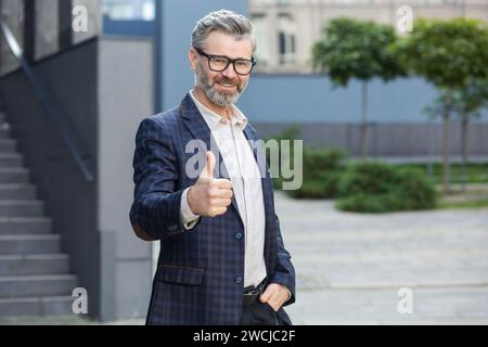 Homme d'affaires mûr souriant dans un costume élégant montrant le geste pouce vers le haut en dehors d'un environnement de bureau contemporain. Banque D'Images