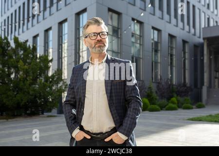 Dirigeant masculin mature regardant confiant dans un costume d'affaires, debout à l'extérieur d'un immeuble de bureaux avec une expression déterminée. Banque D'Images