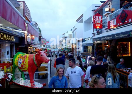 Vacanciers / touristes Walking & Standing sur l'étroite Rua Dr. Frederico Ramos Mendes (rue des bars) dans la soirée à Alvor, Algarve, Portugal, UE Banque D'Images