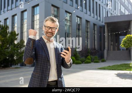 Joyeux cadre dirigeant montrant un geste de victoire avec un poing levé tout en tenant un smartphone, debout à l'extérieur d'un bureau moderne. Banque D'Images
