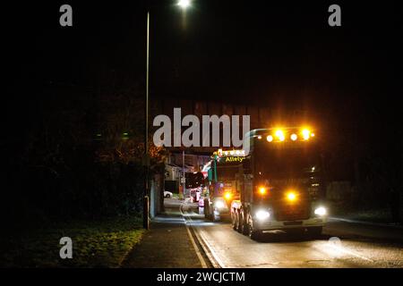 Merthyr Tydfil, Galles du Sud, Royaume-Uni. 15 janvier 2024. Un poids lourd de charge anormale a traversé le centre-ville lundi soir, après avoir été incapable de terminer son voyage à Hirwaun la nuit dernière à cause du mauvais temps. Le camion géant de 78m a été escorté par la police et les routes fermées pour permettre le passage du camion, ce qui a attiré une foule nombreuse qui l'a regardé passer. Crédit : Andrew Bartlett/Alamy Live News Banque D'Images