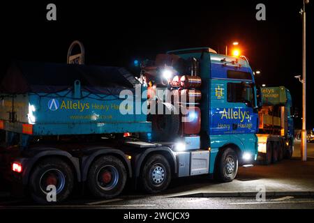 Merthyr Tydfil, Galles du Sud, Royaume-Uni. 15 janvier 2024. Un poids lourd de charge anormale a traversé le centre-ville lundi soir, après avoir été incapable de terminer son voyage à Hirwaun la nuit dernière à cause du mauvais temps. Le camion géant de 78m a été escorté par la police et les routes fermées pour permettre le passage du camion, ce qui a attiré une foule nombreuse qui l'a regardé passer. Crédit : Andrew Bartlett/Alamy Live News Banque D'Images