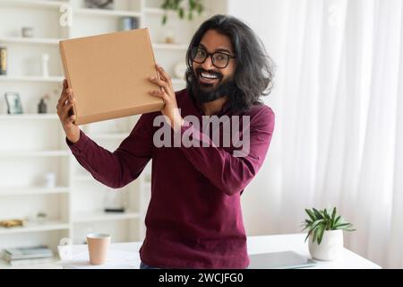Homme indien joyeux tenant une boîte en carton tout en se tenant près du bureau à la maison Banque D'Images