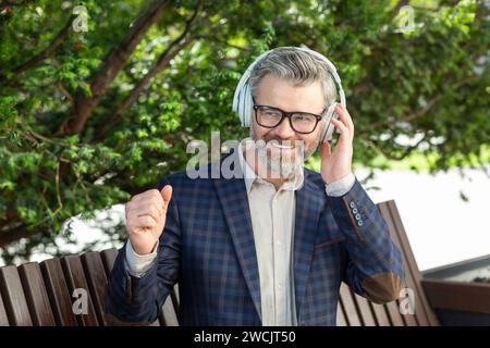 Un homme d'affaires senior souriant dans un costume élégant se détend avec de la musique sur des écouteurs assis à l'extérieur, exsudant la confiance. Banque D'Images