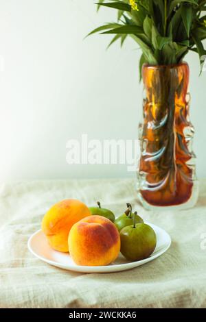 Nature morte avec des pêches fraîches et des prunes sur fond de toile de lin sur la table en bois. Fruits d'été juteux lumineux sur une assiette blanche avec des fleurs jaunes Banque D'Images