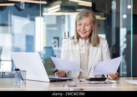 Femme d'affaires senior souriante travaillant dans le bureau au bureau, tenant des documents dans les mains, examinant l'affaire et les factures. Banque D'Images