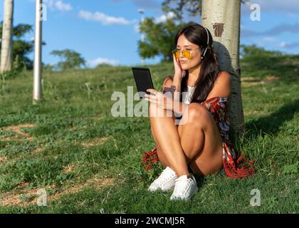 Femme détendue avec des lunettes de soleil en utilisant des écouteurs tout en lisant un ebook assis à l'extérieur dans un parc. Concept technologique. Banque D'Images