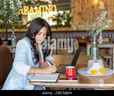 Femme écrivant sur un cahier tout en travaillant avec un ordinateur portable dans un café. Technologie et concept commercial. Banque D'Images