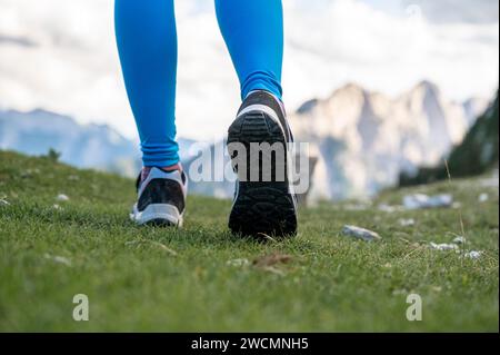 Vue à angle bas des jambes féminines dans les chaussures de randonnée marchant sur l'herbe verte avec de hautes montagnes en arrière-plan. Vue de la semelle des chaussures. Banque D'Images