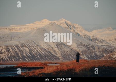 Pic islandais enneigé surplombant une rivière sinueuse, encadré par un paysage serein. Une figure solitaire en noir absorbe la vue à couper le souffle. Banque D'Images