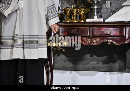 Un acolyte, vêtu de sa robe blanche, tient une cloche dans sa main gauche, debout à côté d'une table avec plusieurs ambules dorées. Banque D'Images