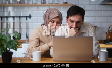 Couple nouvellement marié assis dans la cuisine en utilisant un ordinateur portable attristé par les nouvelles, la perte, la femme console le mari. Déçu mauvaise nouvelle, perte ne Banque D'Images