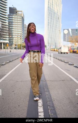 Une jeune femme joyeuse dans un col roulé violet rit en marchant dans une rue urbaine Banque D'Images
