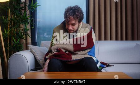 Homme de cheveux bouclés malade se couvrant avec une couverture chaude, souffrent de toux et d'éternuements de nez qui coule tout en étant assis sur un canapé à la maison moderne Banque D'Images