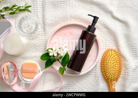 Bouteille à pompe non étiquetée placée sur un plateau en marbre avec des fleurs blanches. Une brosse, un coussin compact, une bougie parfumée et un verre d'eau affichés. Nettoyant pro Banque D'Images