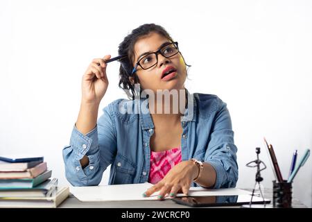 Une jolie jeune étudiante indienne en lunettes pensant tout en étudiant sur la table d'étude et fond blanc Banque D'Images