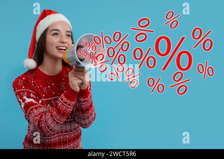 Offre de réduction. Femme en pull de Noël et chapeau de Père Noël criant en mégaphone sur fond bleu clair. Signes de pourcentage sortant de l'appareil Banque D'Images