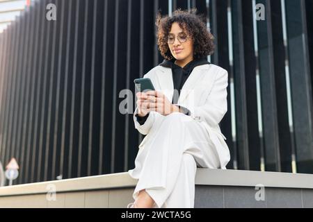 Une jeune femme professionnelle confiante vêtue d'un élégant costume blanc est assise à l'extérieur d'un bâtiment moderne tout en utilisant son smartphone Banque D'Images
