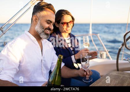 Couple heureux assis à la table en bois et versant du champagne dans des verres à vin tout en profitant du temps sur le yacht Banque D'Images