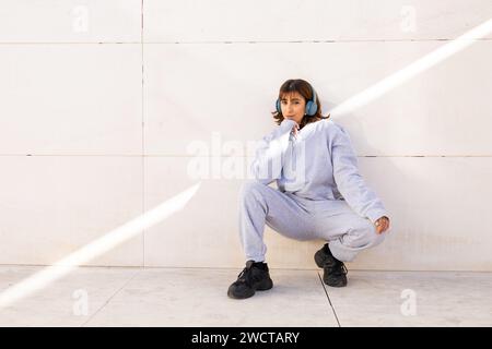 Une jeune femme dans un survêtement bleu confortable et des baskets noires s'assoit sur un fond minimaliste, écoutant de la musique avec des écouteurs bleus Banque D'Images