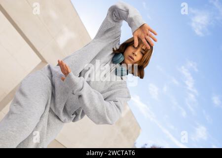 Une jeune femme prend une pose élégante dans son survêtement gris confortable, accessoirisé avec des écouteurs sans fil tendance, sous un ciel bleu clair Banque D'Images