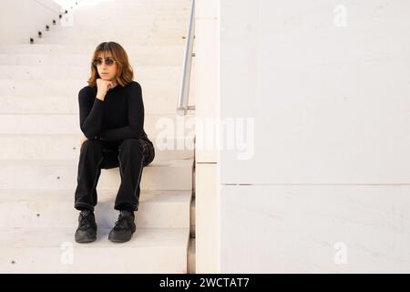 Une jeune femme contemplative portant des lunettes de soleil et une tenue noire est assise seule sur des escaliers blancs, exsudant un sentiment de style urbain décontracté et d'introspection Banque D'Images