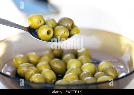 Une cuillerée d'olives vertes dénoyautées est maintenue au-dessus d'un bol en verre rempli d'un assortiment similaire sur un fond neutre Banque D'Images