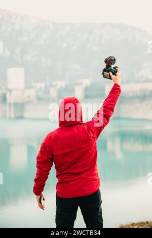 Vue arrière personne dans un sweat à capuche rouge prenant un selfie avec une caméra devant un lac tranquille et paysage de barrage Banque D'Images