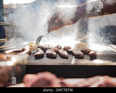 Recadrée mains de chef masculin adulte anonyme tout en se tenant près de la grille de barbecue couvert par la fumée avec des saucisses à la lumière du jour et en utilisant la fourchette en acier à TUR Banque D'Images
