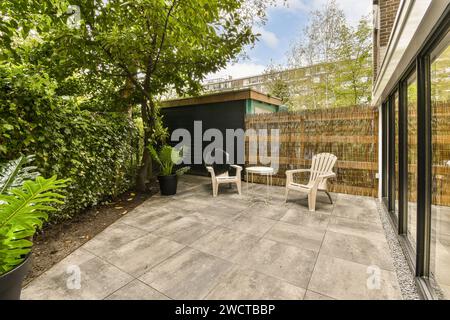 Une cour arrière urbaine sereine présente un patio avec des chaises pour la détente, entouré de verdure luxuriante et une clôture en bambou pour la vie privée. Banque D'Images