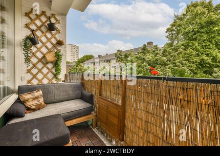 Un balcon urbain accueillant avec un canapé confortable, un banc de rangement en bois et un treillis avec des plantes suspendues sur fond de verdure. Banque D'Images