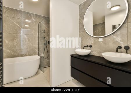 Salle de bain contemporaine avec carrelage mural en marbre poli, cabine de douche en verre sans cadre, baignoire sur pied, vasques ovales sur meuble sombre Banque D'Images